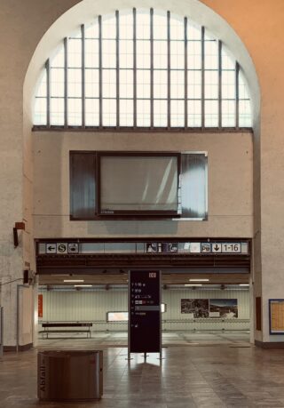 Großzügiger Bahnhofsinnenraum mit großem Bogenfenster, beleuchtetem Wegweiser, leeren Bänken und Wegweisern über den Bahnsteigeingängen.