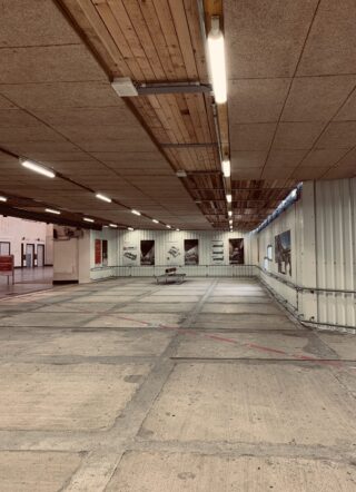 Großer, leerer Innenraum mit Betonboden, Wellblechwänden und fluoreszierender Deckenbeleuchtung; an der gegenüberliegenden Wand hängen Poster und in der Mitte steht eine Bank.