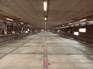 Eine breite, leere Rampe im Innenbereich mit Betonboden und Deckenbeleuchtung, gesäumt von Geländern und Schwarzweißfotos auf beiden Seiten.