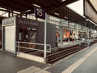 Ein moderner Bahnsteig mit einem geschlossenen Café-Kiosk mit der Aufschrift „Dean & David“ unter dem Schild 15 und einem im Hintergrund sichtbaren Zug.