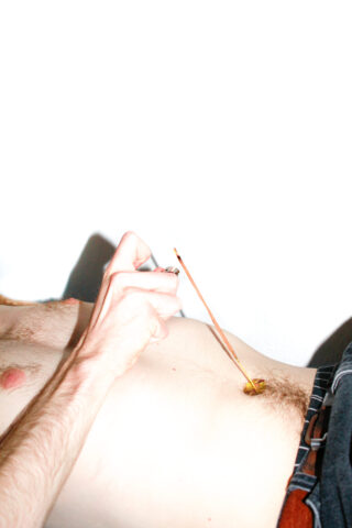 Eine Person liegt ohne Hemd da, hat ein Räucherstäbchen in ihrem Bauchnabel und hält es mit einer Hand fest.