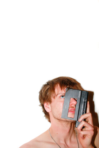 Eine Person ohne Hemd hält einen alten Kassettenrekorder von Sony senkrecht über ein Auge und blickt vor einem schlichten weißen Hintergrund nach oben.