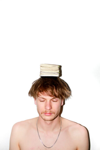 Ein Mann ohne Hemd und mit geschlossenen Augen balanciert einen kleinen Stapel Bücher auf seinem Kopf vor einem schlichten weißen Hintergrund.
