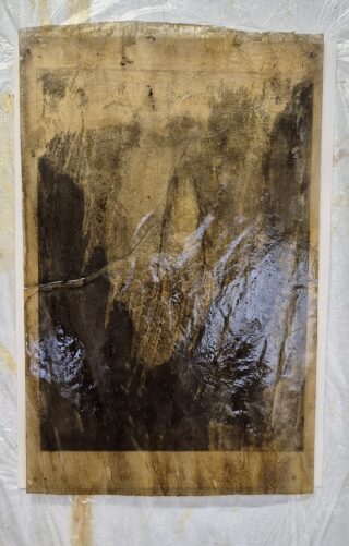 Ein großes, rechteckiges Blatt Papier, bedeckt mit dunklen Flecken und einem klebrigen, glänzenden Rückstand, liegt auf einer zerknitterten, halbtransparenten Oberfläche.