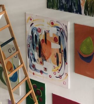 An einer weißen Wand neben einer Holzleiter hängen mehrere abstrakte Gemälde; ein Gemälde zeigt farbenfrohe, wirbelnde Formen und Linien.