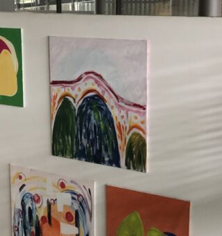 An einer weißen Wand hängen drei abstrakte Gemälde; das mittlere Stück zeigt Bogenformen und bunte Pinselstriche in Grün-, Blau- und Rottönen.