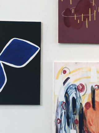 An einer weißen Wand hängen drei abstrakte Gemälde; eines zeigt eine blaue Form, ein anderes kastanienbraune Formen und Linien und das dritte enthält bunte Pinselstriche mit roten Kreisen.