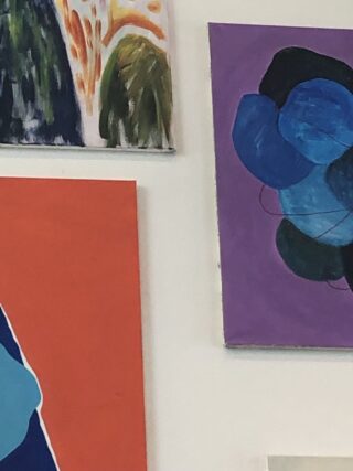 An einer weißen Wand hängen drei abstrakte Gemälde; eines ist in Grün und Orange gehalten, ein anderes in Rot und Blau und das dritte zeigt blaue und schwarze Formen auf violettem Hintergrund.