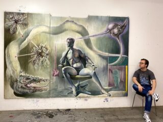 Eine Person sitzt auf einem Stuhl neben einem großen surrealen Gemälde, das auf einer vierteiligen Leinwand eine sitzende Figur zeigt, die von abstrakten Kreaturen und Formen umgeben ist.