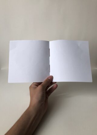 Eine Hand hält vor einem schlichten, hellen Hintergrund ein kleines, leeres, weißes Heft aufgeschlagen.