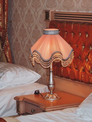 Eine dekorative Tischlampe mit einem Stoffschirm mit Fransen steht auf einem Nachttisch aus Holz zwischen zwei Betten mit weißen Kissen in einem reich verzierten Schlafzimmer.