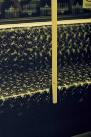 Gemusterte Sitzgelegenheiten in einer U-Bahn oder einem Zug mit einer gelben Stange in der Mitte; im Fenster sind verschwommene Bahnhofsgrafiken zu sehen.