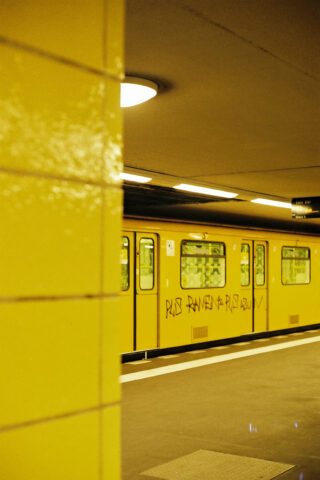 Eine gelbe U-Bahn mit Graffiti hält an einer U-Bahn-Station mit gelb gefliesten Wänden und Deckenbeleuchtung.