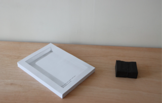 Ein dickes weißes Buch mit einem grauen Einband liegt auf einer hellen Holzoberfläche neben einem kleinen, gefalteten schwarzen Gegenstand.