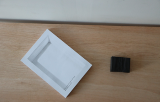 Ein weißer rechteckiger Gegenstand, möglicherweise ein Buch, wird diagonal auf einer hellen Holzoberfläche neben einem kleinen, schwarzen rechteckigen Gegenstand platziert.
