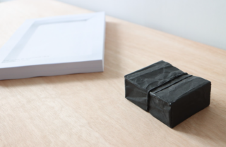 Ein Stapel weißer Papiere und zwei kleine, schwarze, zerknitterte rechteckige Objekte liegen auf einer hellen Holzoberfläche.