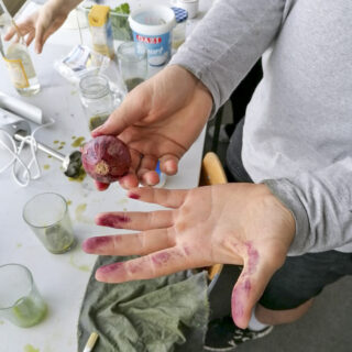 Eine Person hält eine rote Zwiebel und zeigt lila gefärbte Finger in einer Küchenumgebung mit verschiedenen Lebensmitteln und Utensilien auf dem Tisch.