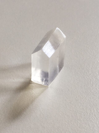 Ein klarer, prismenförmiger Kristall mit einer schrägen Spitze auf einer glatten, hellen Oberfläche, der einen schwachen Schatten wirft.