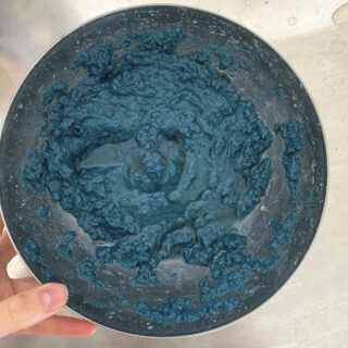 Eine Hand hält eine Metallschüssel mit einer dicken, klumpigen blauen Mischung auf einer Edelstahloberfläche.