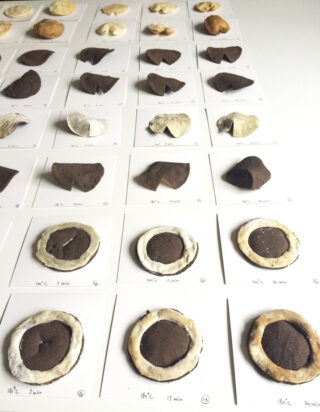 Reihen von braunen und weißen Tonproben sind auf beschrifteten weißen Karten angeordnet, wobei auf jeder Karte unterschiedliche Temperaturen und Zeiten angegeben sind und wahrscheinlich ein Brenntest dokumentiert wird.