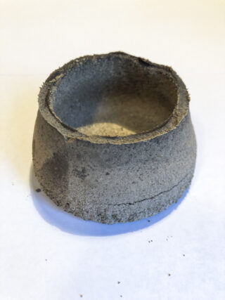 Auf einer weißen Fläche steht ein grob geformter, grauer, unbearbeiteter Tontopf mit weiter Öffnung und unebenen Rändern.