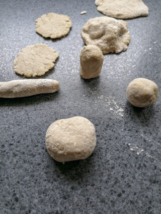 Verschiedene Formen aus rohem Teig, darunter eine Kugel, eine Scheibe und eine zylindrische Form, sind auf einer grauen, strukturierten, mit Mehl bestäubten Oberfläche angeordnet.