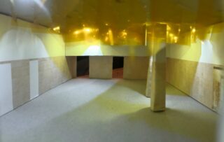 Ein Raummodell mit beigen Wänden, hellgrauem Boden, rechteckigen Säulen und einer gelb reflektierenden Decke, von einem Ende des Raums aus betrachtet.