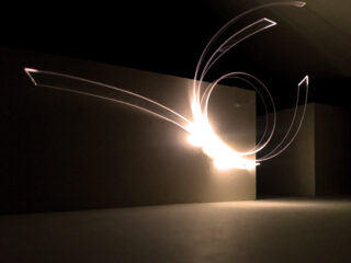 Ein Foto mit Langzeitbelichtung zeigt weiße Lichtspuren, die vor einem schlichten, dunklen Hintergrund mit rechteckigen Strukturen geschwungene Muster bilden.