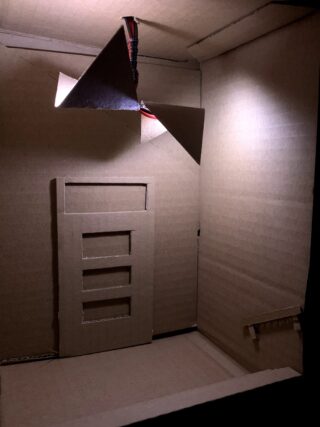 Ein kleiner Kartonraum verfügt über eine Tür mit drei Fensterausschnitten, eine Treppe und eine Deckenleuchte aus gefaltetem Karton mit daran befestigten Drähten.