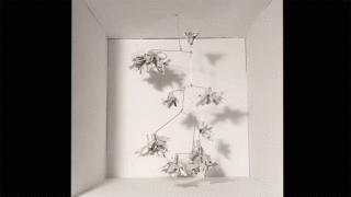 Ein weißes, skulpturales Mobile aus papierähnlichen Formen hängt in einer schlichten weißen Box und dreht sich langsam. Schatten des Mobiles erscheinen an Wänden und Boden.