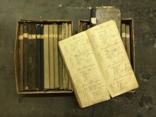 Zwei Kisten mit alten, gebundenen Hauptbüchern auf einem Betonboden. In einem offenen Hauptbuch sind handschriftliche Einträge mit Tinte zu sehen.