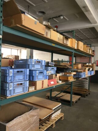In Metallregalen in einem Lagerhaus werden Kartons, blaue Kunststoffkisten und Stapel aus Holz oder Brettern aufbewahrt; einige Artikel liegen auf Holzpaletten auf dem Boden.