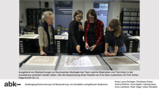 Vier Personen stehen um einen Tisch herum und betrachten ein Kunstwerk unter hellem Licht und diskutieren über die Materialien und Techniken in einer Atelierumgebung.