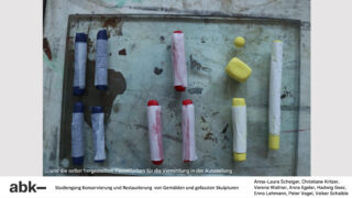 Verschiedene Pastellstifte, teilweise in Papier eingewickelt, sind auf einer Glasfläche angeordnet. Am unteren Bildrand sind teilweise deutscher Text und Bildnachweise zu sehen.