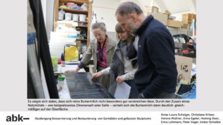 Drei Personen untersuchen ein dunkles, rechteckiges Objekt in einer Werkstatt voller Künstlerbedarf und Lagerregale. Text und Bildnachweis erscheinen unter dem Bild.