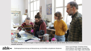 Vier Personen stehen in einem Atelier um einen mit Künstlerbedarf und Papieren bedeckten Tisch herum und besprechen Restaurierungsarbeiten. Eine Person gestikuliert, während die anderen zuschauen.