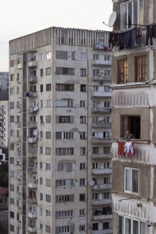 Zwei alte Wohnhäuser mit sichtbaren Balkonen, an manchen hängt Wäsche draußen; auf einem Balkon steht eine Person, über deren Geländer Decken hängen.