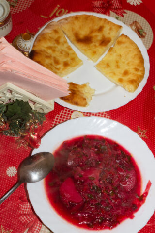 Ein Teller mit geschnittenem Brot und eine Schüssel Rote-Bete-Suppe mit Kräutern stehen auf einer roten Festtischdecke, daneben liegen ein Löffel und Servietten.