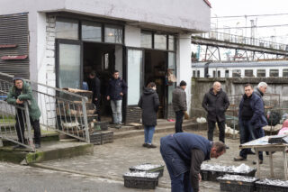 Vor einem Gebäude stehen und gehen Menschen umher, in der Nähe von Stapeln voller Fischkisten. Im Hintergrund ist ein Zug auf den Gleisen zu sehen.