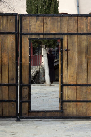 Ein Holztor mit schmalem Türausschnitt steht offen und gibt den Blick auf eine schwarz gekleidete Person frei, die im Hintergrund bei einem Baum steht.
