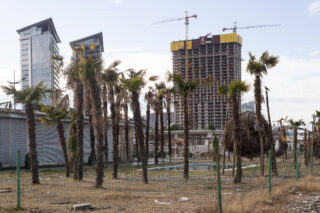 Ein eingezäuntes Gelände mit spärlichen, trockenen Palmen vor einem ungenutzten Pool und im Hintergrund im Bau befindlichen Hochhäusern.