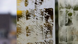 Nahaufnahme von zwei Milchglasplatten mit abstrakten, strukturierten Mustern in Gelb- und Grüntönen, Licht und Gebäude im Hintergrund unscharf.