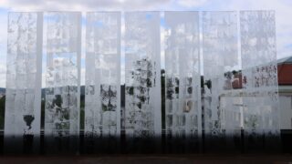 Sieben Milchglasplatten mit strukturierten, halbtransparenten Mustern sind vertikal vor dem Hintergrund von Gebäuden und bewölktem Himmel angeordnet.