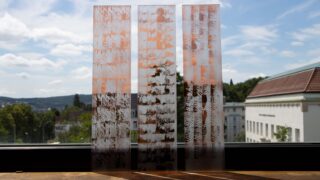 Drei rechteckige Glasplatten mit strukturierten, rostigen Mustern stehen aufrecht auf einer Fensterbank, mit Blick auf Gebäude, Bäume und einen bewölkten Himmel im Hintergrund.