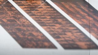 Schatten eines gemusterten Objekts mit diagonalen Linien auf einer hellen Oberfläche, wodurch abwechselnd orange und weiße Abschnitte entstehen.