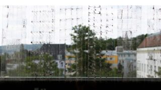 Ein verschmiertes, schmutziges Glasfenster mit vertikalen Streifen und Flecken verdeckt teilweise die Sicht auf Gebäude, Bäume und einen bewölkten Himmel draußen.