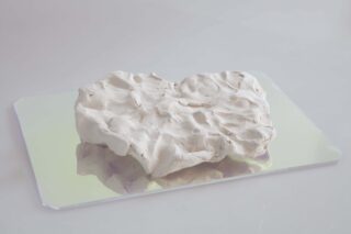 Unregelmäßig geformtes Stück aus weißem Ton mit strukturierter Oberfläche, das auf einem rechteckigen Spiegel oder einer reflektierenden Oberfläche ruht.
