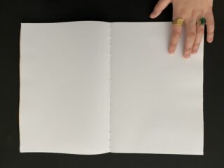 Eine Hand mit Ringen hält ein leeres, weißes, genähtes Notizbuch auf einer schwarzen Oberfläche offen.