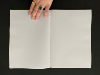 Eine Hand mit zwei Ringen hält vor einem schwarzen Hintergrund ein leeres, weißes Notizbuch mit Spiralbindung offen.