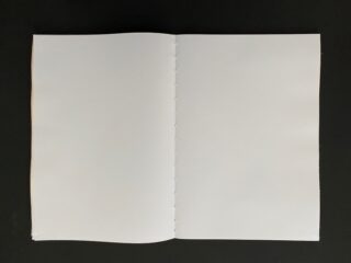 Ein offenes, leeres Buch liegt flach auf einer schwarzen Oberfläche und zeigt zwei leere weiße Seiten mit sichtbaren Bindungslöchern in der Mitte.